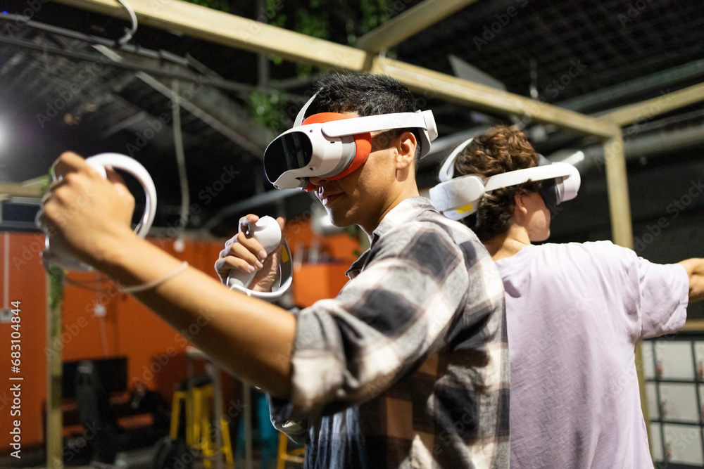 Side view of two friends stand in a gaming area fully immersed in a virtual reality world wearing VR headsets and holding motion controllers Side view of two friends stand in a gaming area fully immersed in a virtual reality world wearing VR headsets and holding motion controllers