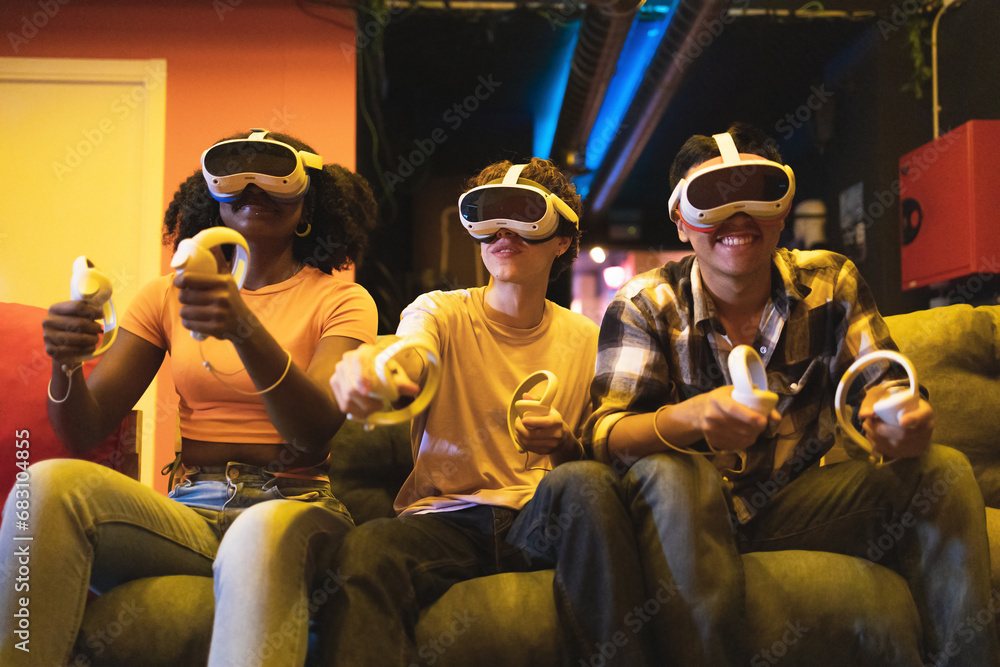 Multiethnic friend engrossed in a virtual reality game wearing VR headsets and holding controllers sitting on a couch in a dimly lit room Multiethnic friend engrossed in a virtual reality game wearing VR headsets and holding controllers sitting on a couch in a dimly lit room