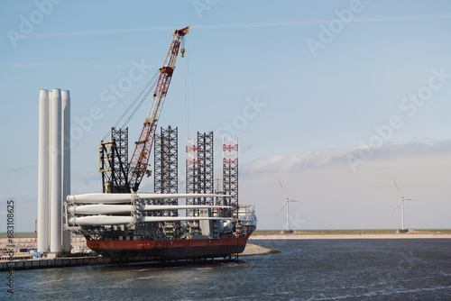Self Elevating Wind Installation Vessel In The European Port During Process Of Heavy Lift Operation And Loading Of The Wind Blades On Deck. Offshore Support Construction DP Vessel Alongside