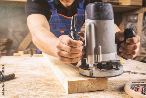  A young brunette man carpenter equals a wooden plank with a milling machine in the workshop