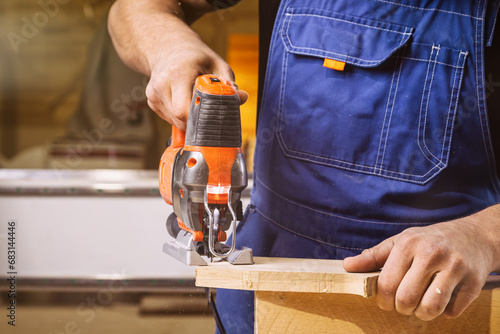 Close up of experienced carpenter in work clothes and small buiness owner  carpenter saw and processes the edges of a wooden bar with a jig saw  in a workshop photo