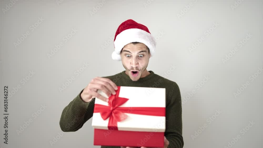 Young hispanic man unpacking gift wearing christmas hat over isolated white background Young hispanic man unpacking gift wearing christmas hat over isolated white background