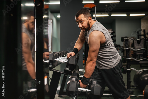 A bearded male bodybuilder takes a dumbbell in his hand and demonstrates the triceps. Work on your body in the gym.