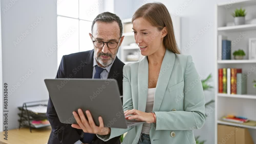 Two confident business workers smiling as they work together on a laptop, standing in the heart of a bustling office environment Two confident business workers smiling as they work together on a laptop, standing in the heart of a bustling office environment