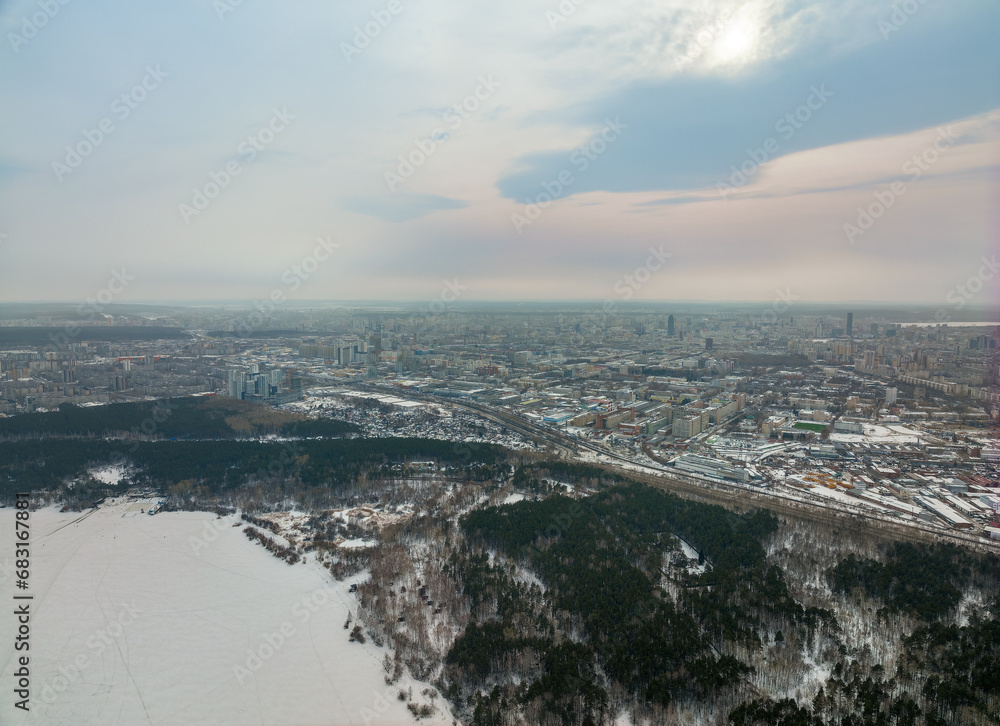 Snow-covered forest on lake shore with ice at sunset and the city on horizon, aerial view