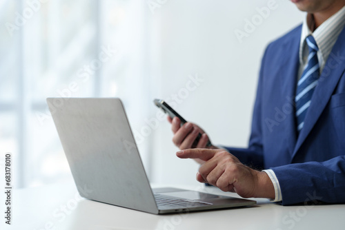 Businessman holds smartphone, checks email, checks job details on Laptop computer on white desk to point to successful business goals in office.
