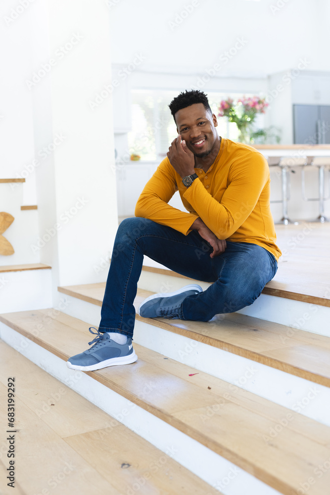 Happy african american man sitting on stairs and scratching face at home, copy space