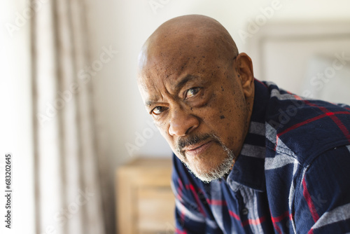 Portrait of thoughtful african american senior man sitting on bed in sunny bedroom