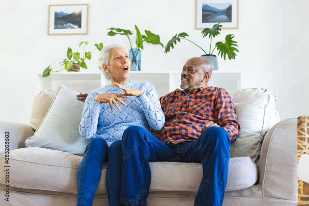 Happy diverse senior couple sitting on sofa, talking and embracing in sunny living room Happy diverse senior couple sitting on sofa, talking and embracing in sunny living room