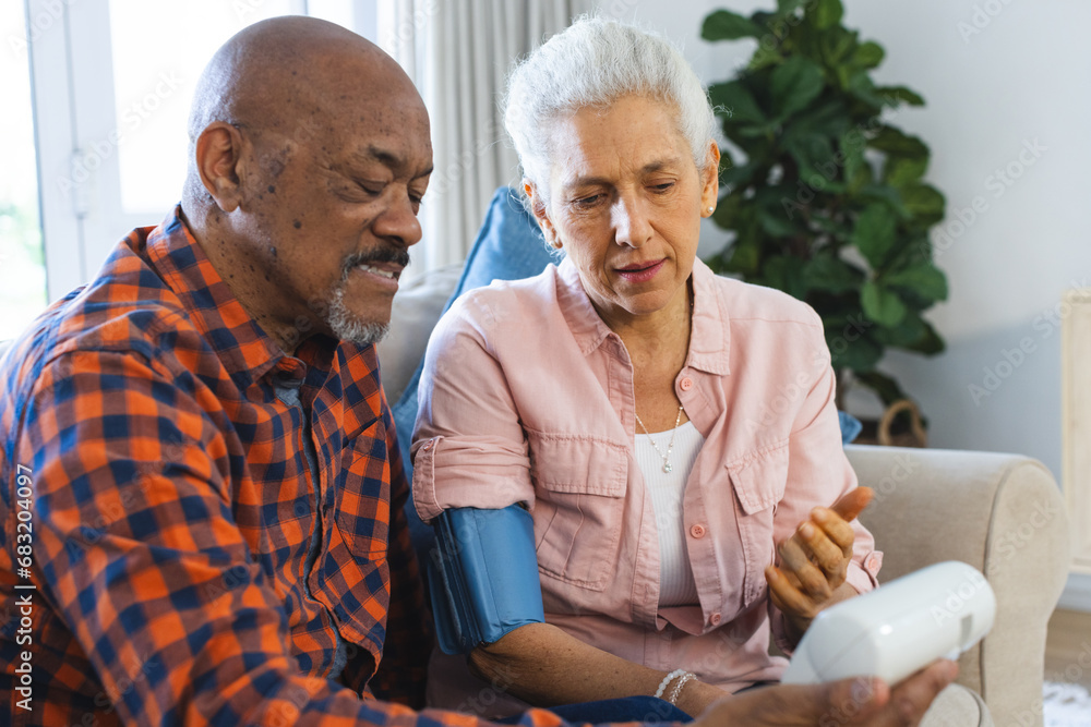 Diverse senior man testing blood pressure of senior woman in sunny living room Diverse senior man testing blood pressure of senior woman in sunny living room