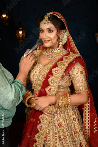 Stunning Indian bride dressed in traditional bridal lehenga with heavy gold jewellery and veil posing fashionable with makeup brush in studio lighting. Wedding fashion and lifestyle.