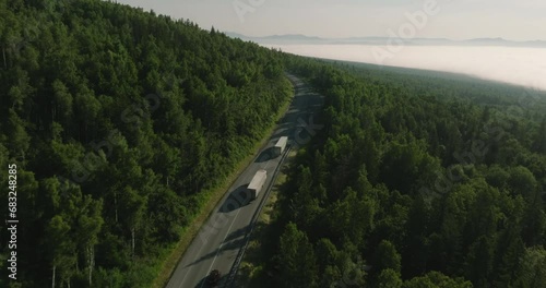 Two Semi Trucks with white trailer and cab driving, traveling alone on dense forest mountain winding road, highway perspective view follow vehicle aerial footage. Freeway trucks traffic
