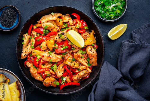 Stir fry chicken with pineapple, red paprika, chives, soy sauce and sesame seeds in frying pan. Asian cuisine dish. Black table background, top view