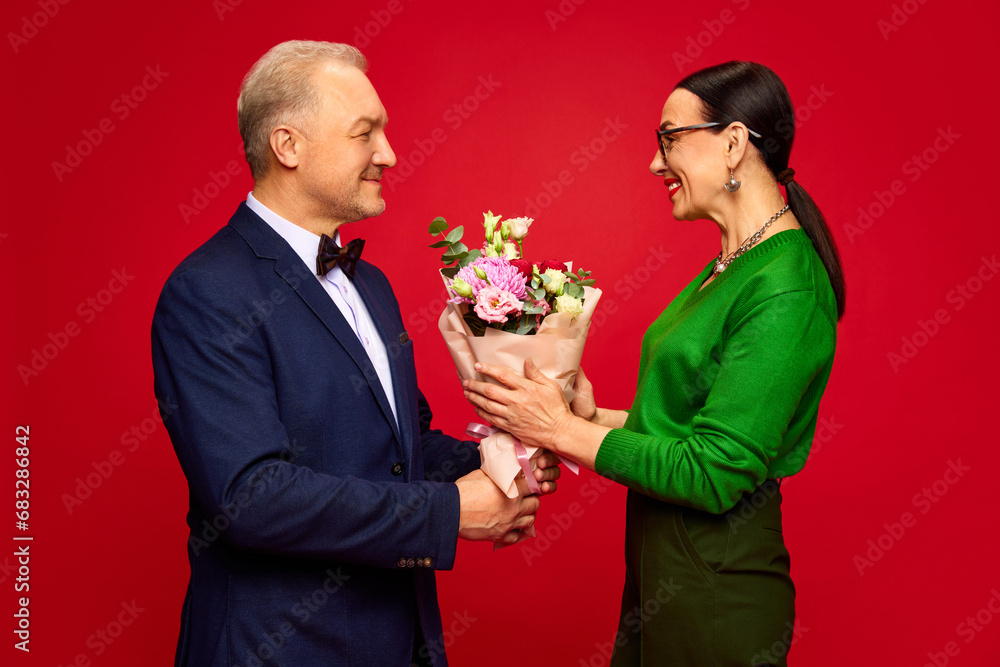 Going on a romantic date. Senior man presenting flower to his wife, celebrating holiday against red studio background. Concept of marriage, relationship, Valentine's Day, love, emotions, fashion Going on a romantic date. Senior man presenting flower to his wife, celebrating holiday against red studio background. Concept of marriage, relationship, Valentine's Day, love, emotions, fashion