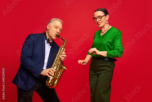 Caring, loving man, husbands playing saxophone to his beautiful wife against red studio background. Romantic date, holiday. Concept of marriage, relationship, Valentine's Day, love, emotions, fashion