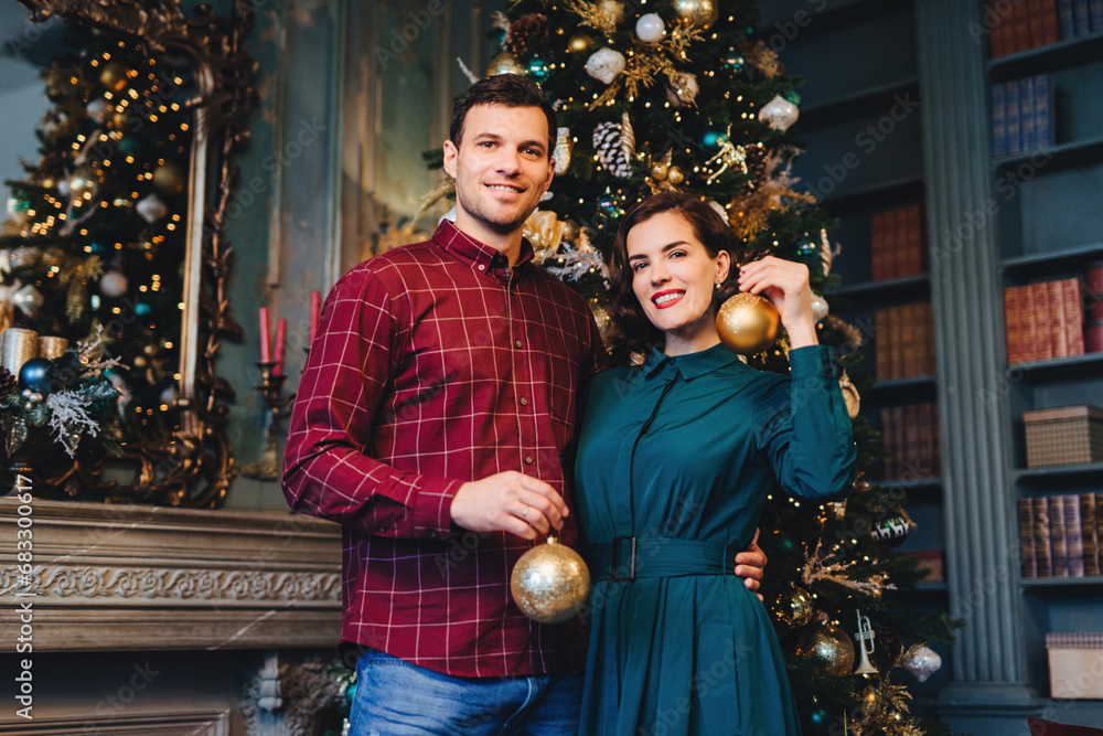 Couple in festive attire holding golden Christmas ornaments, standing by a richly decorated tree in an elegant room.