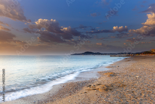 Beautiful sandy - pebbly long beach on the island of Rhodes at sunrise. White waves crash into the shore with a blue sky in the background.