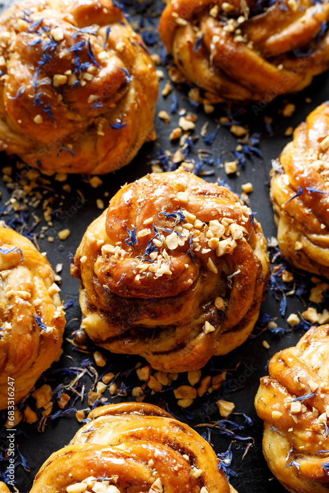 Sweet cardamom rolls sprinkled with chopped nuts and dried cornflower petals on a dark background, focus on the bun inside, top view. Homemade sweet pastries