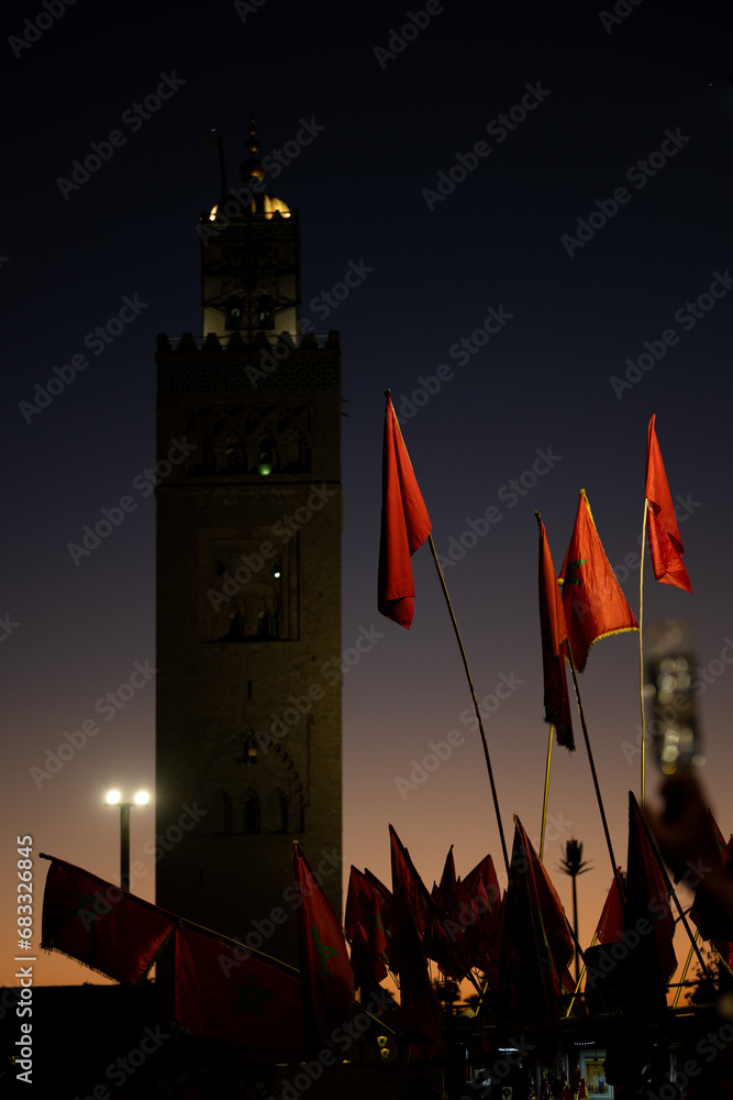 (Selective focus) Stunning view of some Moroccan flags in the foreground and defocused Kutubiyya Mosque in the background at sunset. Marrakesh, Morocco.
