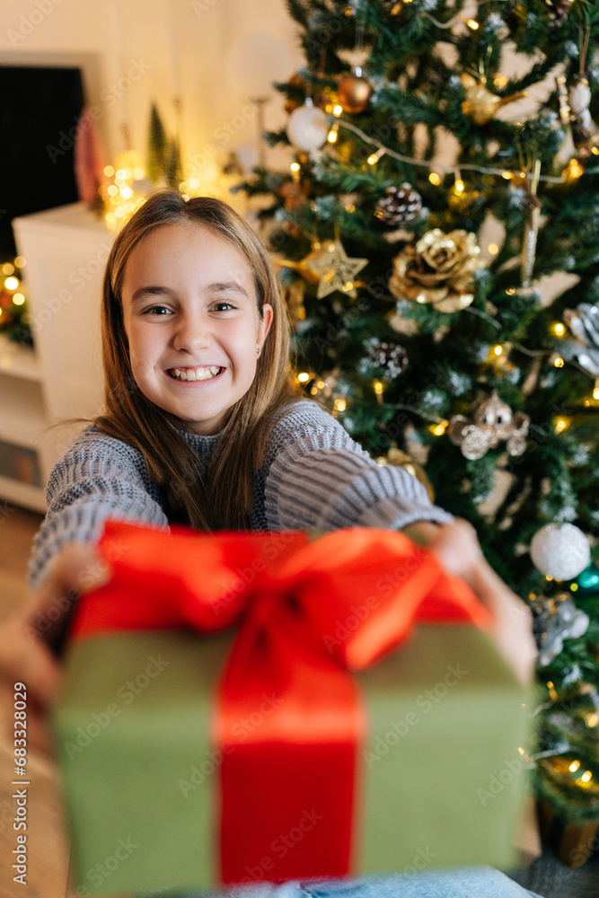 Vertical portrait of cheerful cute little girl holding, giving and showing at camera wrapped with red ribbon and bow festive Christmas present gift box, sitting by glowing xmas tree, looking at camera Vertical portrait of cheerful cute little girl holding, giving and showing at camera wrapped with red ribbon and bow festive Christmas present gift box, sitting by glowing xmas tree, looking at camera