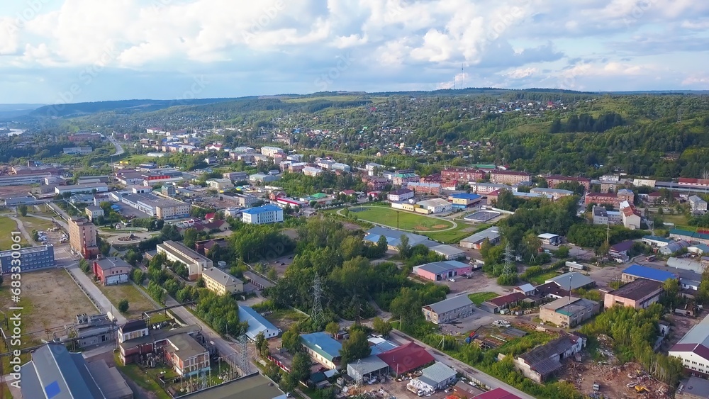 Top view of town with green forest and hills. Clip. Modern town with infrastructure in hilly forest area. Beautiful landscape of town with hilly horizon on summer day