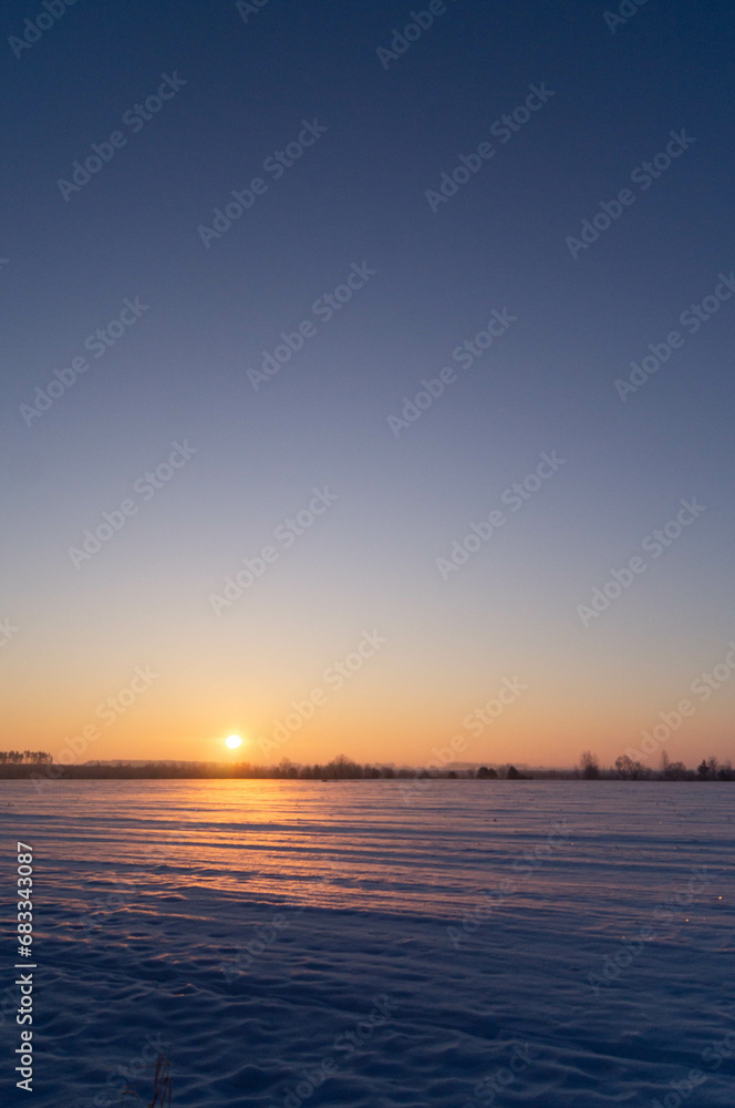 Golden sunrise introduces the day over a glistening, tranquil snowscape, bringing life to a serene winter morning as soft sun rays embrace the frosty, snow-draped landscape with warmth and promise.