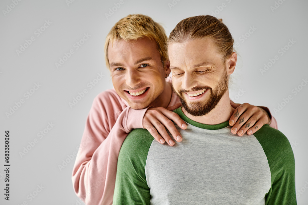 happy gay man in casual attire hugging shoulders of bearded boyfriend and looking at camera on grey happy gay man in casual attire hugging shoulders of bearded boyfriend and looking at camera on grey