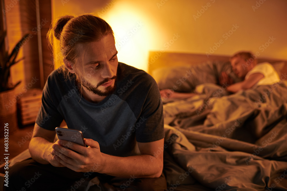 disloyal bearded gay man browsing date app on smartphone near partner sleeping at night in bedroom disloyal bearded gay man browsing date app on smartphone near partner sleeping at night in bedroom