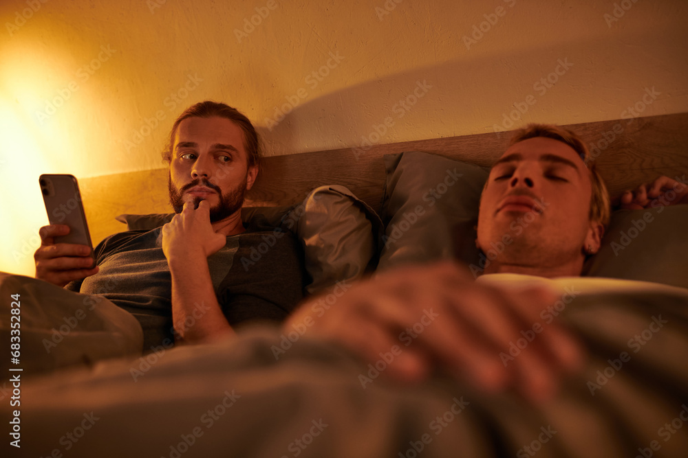 unfaithful bearded gay browsing date app on smartphone near sleeping boyfriend at night in bedroom