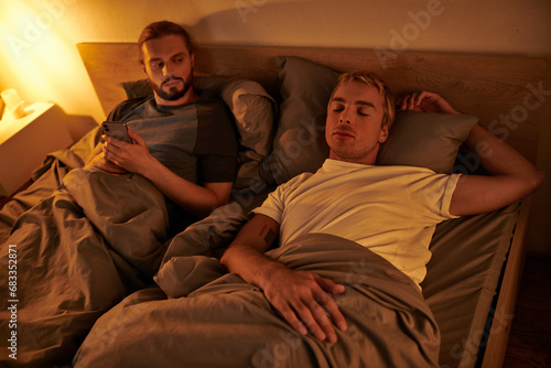 unfaithful bearded gay messaging on mobile phone near sleeping boyfriend at night in bedroom