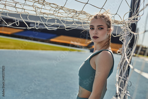 blonde sportswoman with ponytail standing near net after workout in stadium, urban fitness