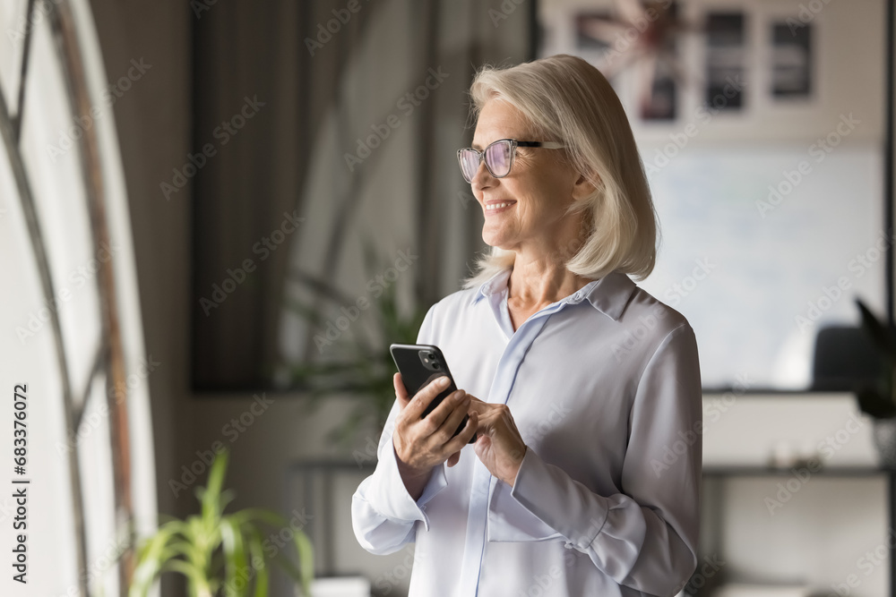 Cheerful elderly business professional woman in glasses getting good news on online chat, holding smartphone, using Internet service, healthcare application, wireless technology for job in office