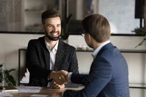 Cheerful young startup leader man shaking hand with investor, business partner at office meeting table. Happy hired job candidate giving handshake with employer, starting career