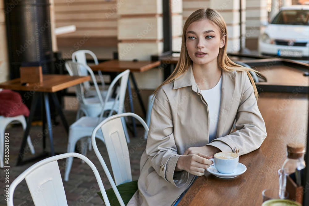 pretty young woman in stylish and beige trench coat looking at her cappuccino in cafe, cup of coffee pretty young woman in stylish and beige trench coat looking at her cappuccino in cafe, cup of coffee