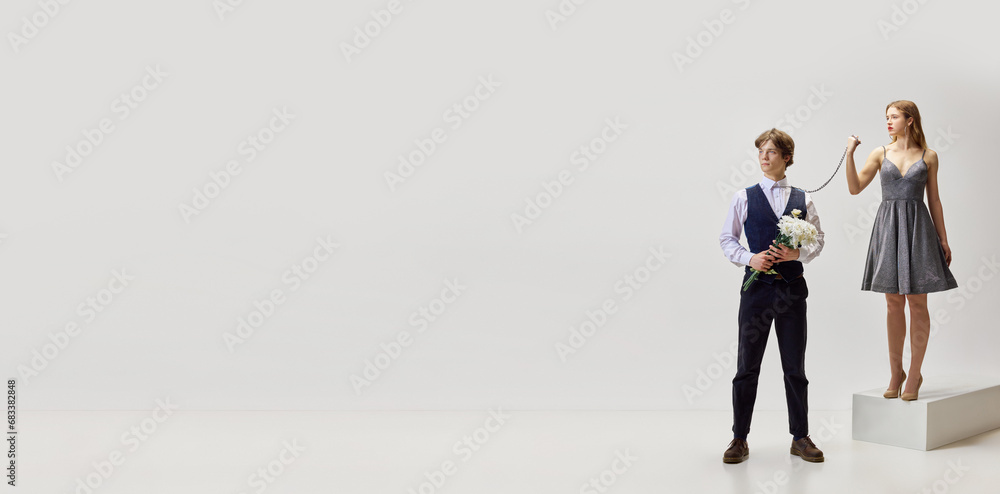 Banner. Modern portrait of young girl tied chain around neck of young man with flower bunch and controlling him against white studio background. Banner. Modern portrait of young girl tied chain around neck of young man with flower bunch and controlling him against white studio background.