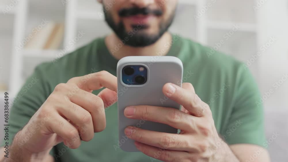 Focus on modern smartphone with violet case in male hands. Blurred background of bearded guy in casual green t-shirt scrolling with finger on smartphone screen while sitting on grey sofa.