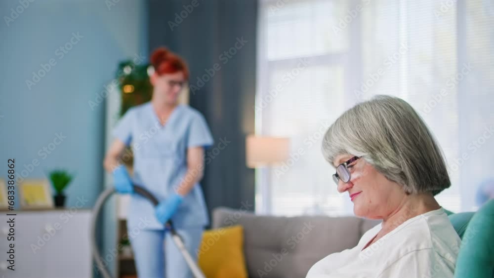 joyful elderly woman reading a book while sitting in a chair while a female social worker helps with cleaning the room joyful elderly woman reading a book while sitting in a chair while a female social worker helps with cleaning the room