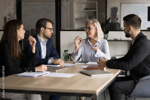 Older mature business team manager woman motivating employees for teamwork, speaking to group of younger colleagues at meeting table, offering ideas for brainstorming, cooperation photo