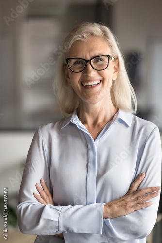 Happy elegant blonde senior businesswoman vertical portrait. Cheerful positive mature business professional woman, CEO, company leader looking at camera with hands crossed ant toothy smile