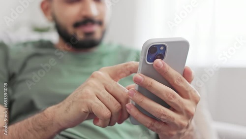 Crop of bearded young man tapping with forefinger on mobile screen while relaxing on comfy couch at home. Hindu male in green t-shirt typing messages while chatting online.