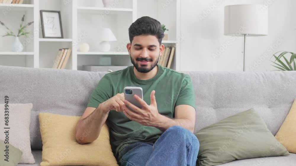 Casually dressed indian man browsing internet on modern smartphone while sitting on grey couch. MIllennial young guy resting at cozy apartment with gadget in hands. Casually dressed indian man browsing internet on modern smartphone while sitting on grey couch. MIllennial young guy resting at cozy apartment with gadget in hands.