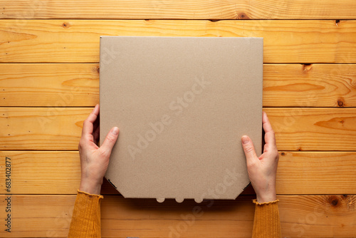 Pizza closed carton box in human caucasian hands on natural wooden table flat lay mockup with blank space