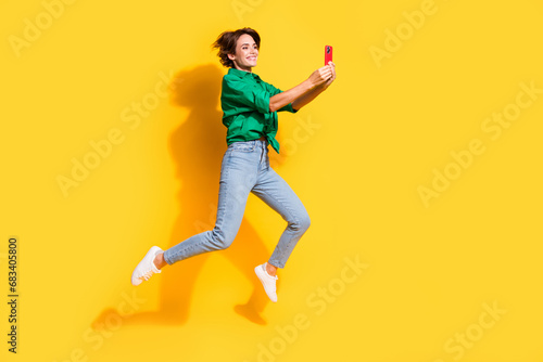 Full length photo of excited pretty girl dressed green shirt jumping chatting modern device isolated yellow color background