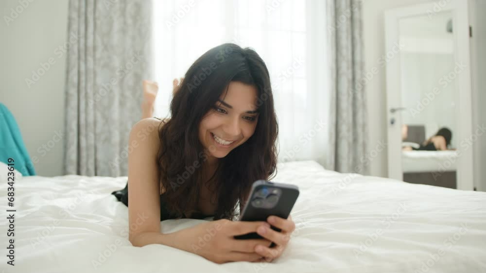 Attractive smiling brunette lying on bed on stomach in bedroom at home typing on phone, chatting. Young woman relaxes, laughs, behaves flirtatiously. High quality 4k footage Attractive smiling brunette lying on bed on stomach in bedroom at home typing on phone, chatting. Young woman relaxes, laughs, behaves flirtatiously. High quality 4k footage