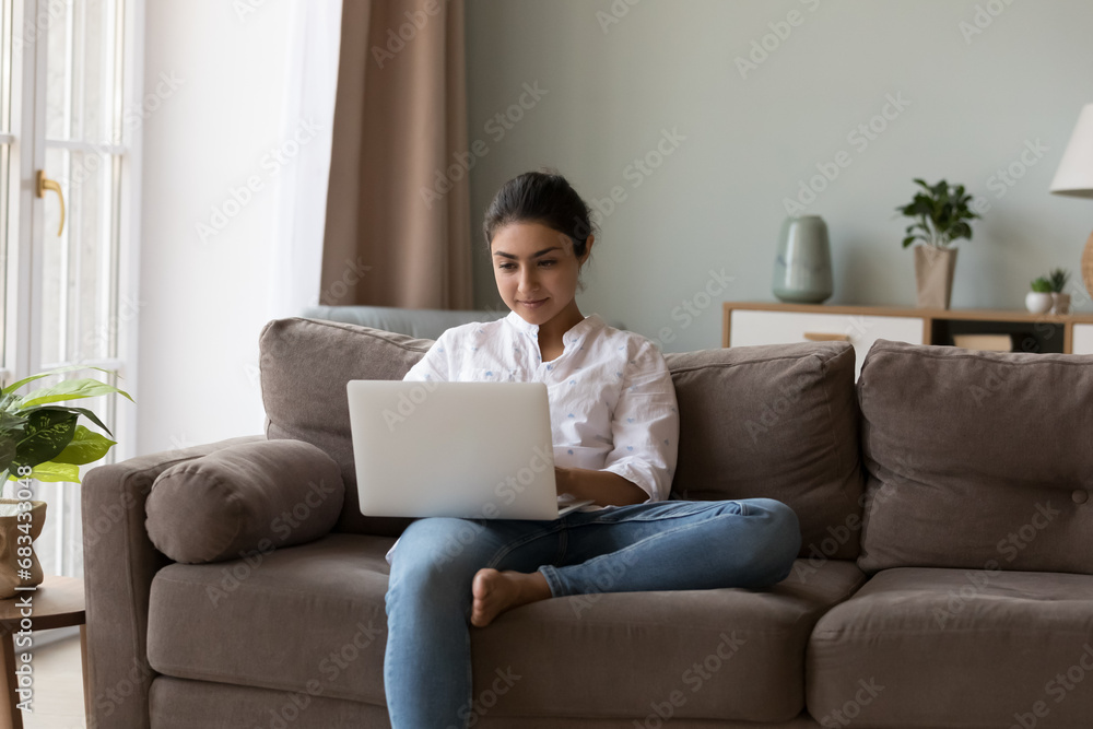 E-commerce, e-shopping, modern technology usage at home concept. Attractive Indian woman sit on sofa using laptop, do telework, looks focused, buying goods on internet, make distancing on-line order E-commerce, e-shopping, modern technology usage at home concept. Attractive Indian woman sit on sofa using laptop, do telework, looks focused, buying goods on internet, make distancing on-line order
