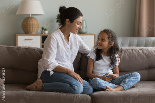 Indian mom and daughter talking sit on sofa. Mum having friendly warm conversation to little girl at home, share experience, give advice, teach child, express love, care. Family communication concept