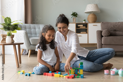 Young loving Indian mother spend weekend leisure with cute little daughter sit on warm floor in living room play multi colored wooden cubes. Children development and growth, fun, games at home concept