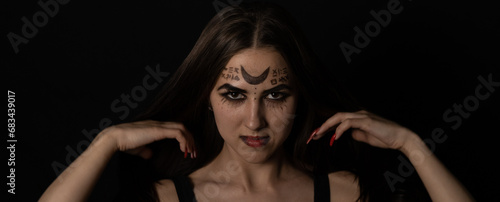 On Halloween night, a witch with red nails straightens her long hair with her hands and stares piercingly into the camera. Porter. A female witch with magical symbols on her face is looking at you. photo