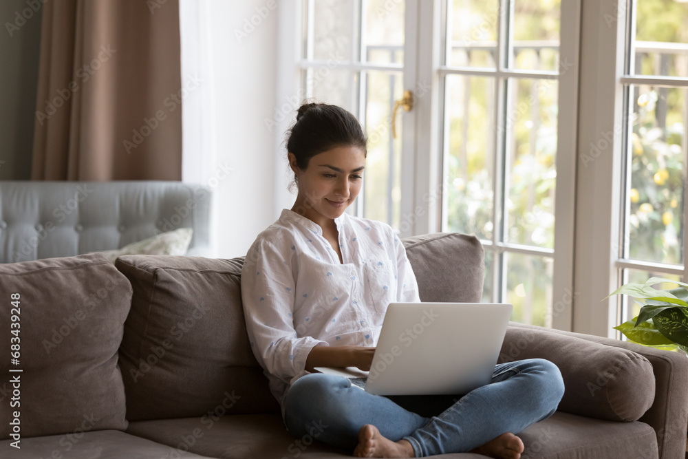 Attractive young Indian woman spend leisure at home using laptop, sit on sofa in living room do e-shopping, busy in distance telework looks focused. E-commerce client, modern tech, lifestyle concept Attractive young Indian woman spend leisure at home using laptop, sit on sofa in living room do e-shopping, busy in distance telework looks focused. E-commerce client, modern tech, lifestyle concept