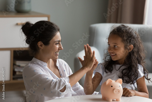 Indian mother and little daughter give high five sit at table with piggy bank. Caring mom teach child be thrifty, save pocket money for future education or purchases, thinking about tomorrow concept